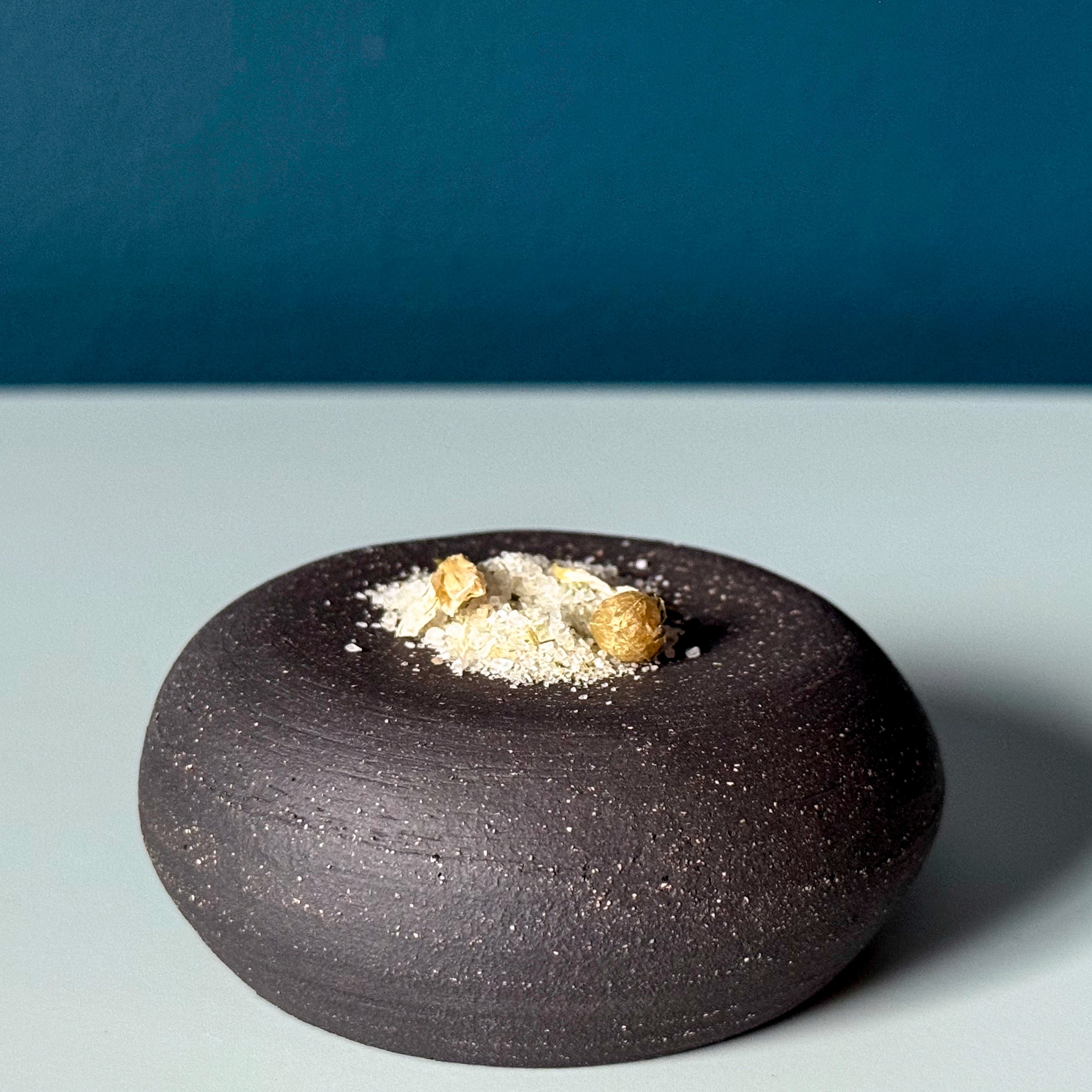Black ceramic bowl with salt flakes and leek flowers on a gray surface with blue background