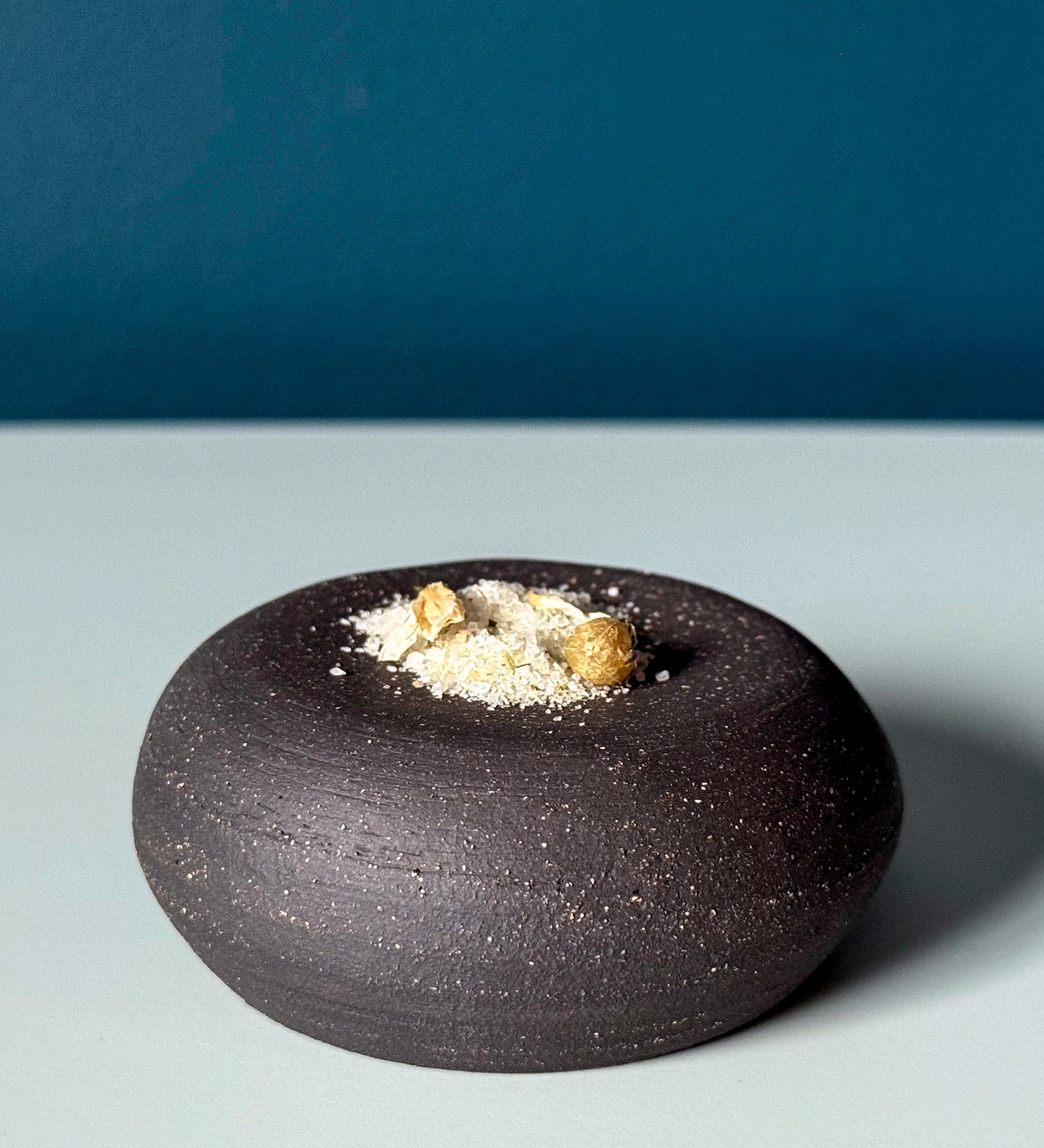 Black ceramic bowl with salt flakes and leek flowers on a gray surface with blue background