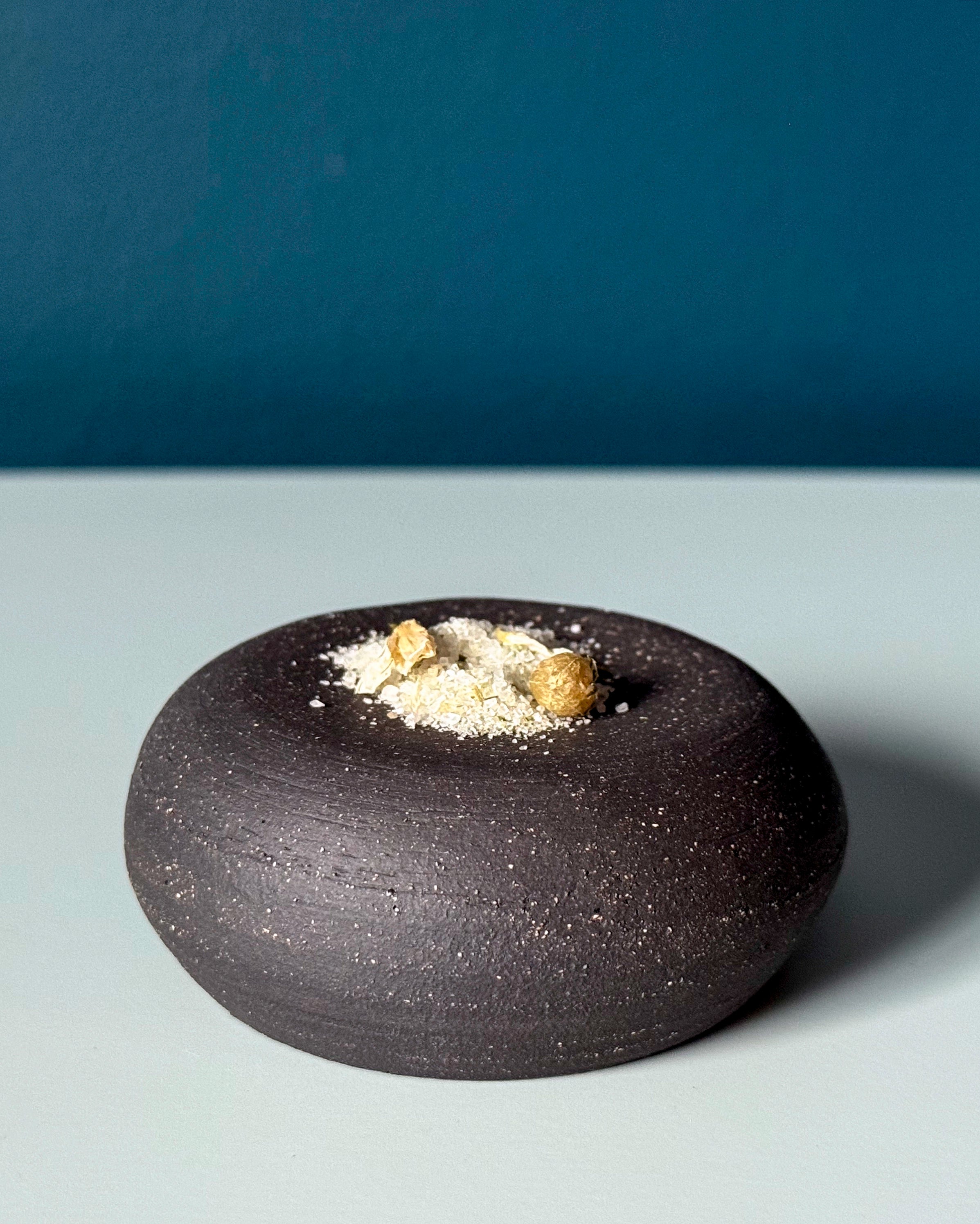 Black ceramic bowl with salt flakes and leek flowers on a gray surface with blue background